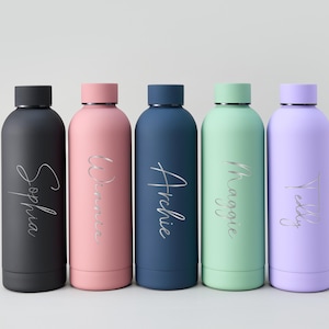May include: Five colorful water bottles in black, pink, navy, mint green, and lavender. Each bottle has a silver name in a cursive font: Sophia, Winnie, Archie, Maggie, and Teddy. The bottles have black or matching colored lids.