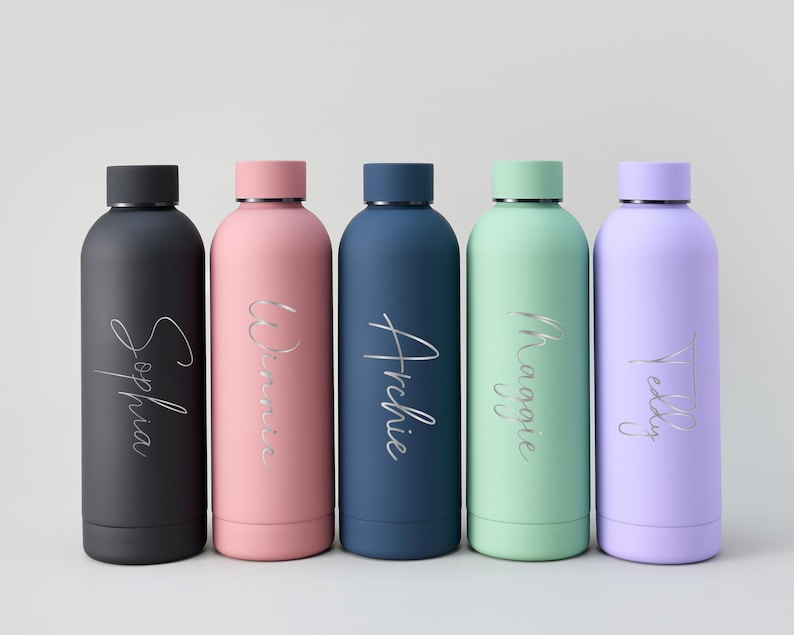 May include: Five colorful water bottles in black, pink, navy, mint green, and lavender. Each bottle has a silver name in a cursive font: Sophia, Winnie, Archie, Maggie, and Teddy. The bottles have black or matching colored lids.