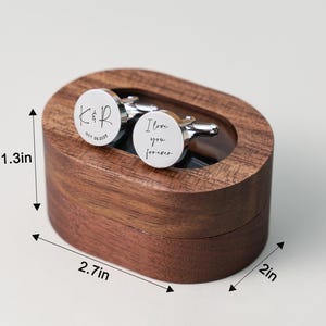 May include: A pair of silver cufflinks with the words "I love you forever" and initials engraved on them, presented in a dark wooden oval box. The box measures 2.7 inches long, 2 inches wide, and 1.3 inches high.