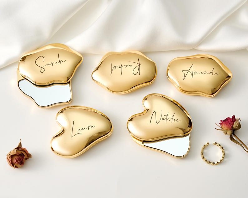 May include: Five gold-colored compact mirrors in various organic shapes, each with a name in elegant script: Sarah, Rachel, Amanda, Laura, and Natalie. Each compact has a mirror. A gold ring and dried rose are also present.