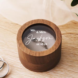 May include: A round, wooden ring box with a clear top, displaying a diamond ring. The box is engraved with "Mr. & Mrs. Jackson" and the date "06.28.24". A second ring is visible in the foreground.