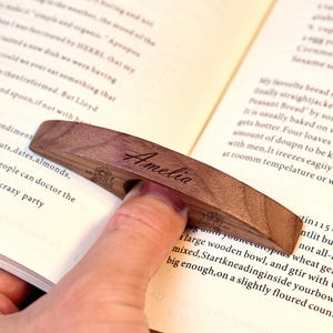 May include: A wooden page holder with the name "Amelia" engraved on it. The page holder is a dark brown color and is being used to hold open a book. The page holder has a curved design.