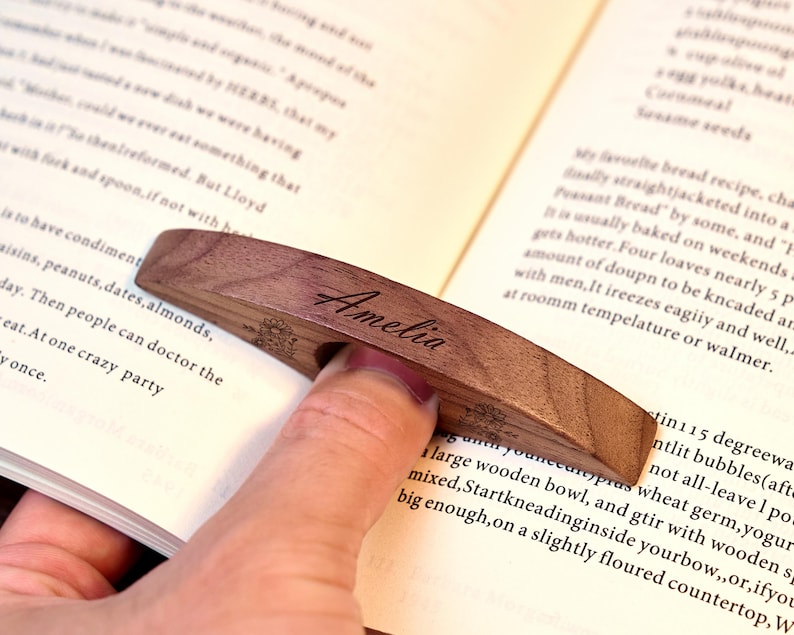 May include: A wooden page holder with the name "Amelia" engraved on it. The page holder is a dark brown color and is being used to hold open a book. The page holder has a curved design.