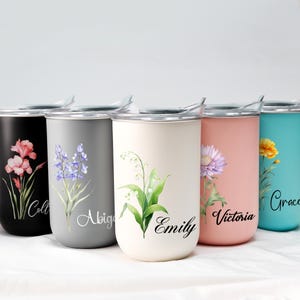 Op de afbeelding: Een set van vijf roestvrijstalen reisbekers in zwart, grijs, cr&egrave;me, roze en teal. Elke beker heeft een bloemenontwerp en een naam in script. De bekers hebben zilveren deksels en zijn ontworpen om dranken warm of koud te houden.