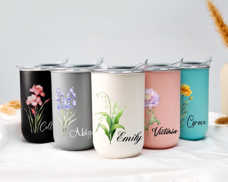 Op de afbeelding: Een set van vijf roestvrijstalen reisbekers in zwart, grijs, cr&egrave;me, roze en teal. Elke beker heeft een bloemenontwerp en een naam in script. De bekers hebben zilveren deksels en zijn ontworpen om dranken warm of koud te houden.
