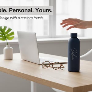 May include: A navy blue water bottle with the name "Sophia" in cursive script. The bottle sits on a light wood desk with a laptop, eyeglasses, and a small vase with a green leaf. Text above reads "Simple. Personal. Yours."