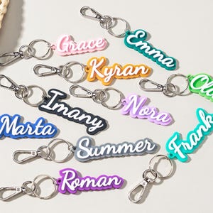 Può includere: Una collezione di portachiavi con nomi colorati. Ogni portachiavi presenta un nome diverso in un carattere giocoso, tra cui "Grace", "Emma" e "Frank". I portachiavi hanno finiture color argento e sono disposti su una superficie chiara.