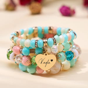 Può includere: Braccialetti impilati con perline in colori pastello, tra cui turchese, rosa e crema. Un ciondolo a forma di cuore dorato con il nome "Kayla" e un cuore è attaccato. I braccialetti presentano varie dimensioni e trame di perline, con accenti dorati.
