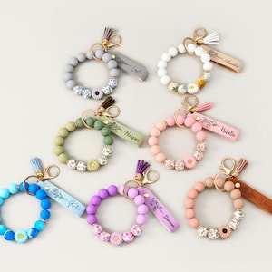 May include: A selection of beaded wristlet keyrings in shades of blue, green, pink, and purple. Each keyring has a tassel and a name tag. Some keyrings feature daisy-shaped beads. Measurements are not available.