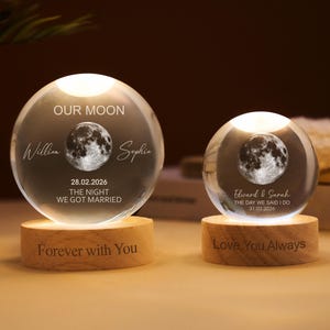 Peut inclure: Deux sphères de cristal éclairées avec un motif de lune, personnalisées avec des noms et des dates. Elles reposent sur des socles en bois, l'un portant l'inscription "Forever with You" et l'autre "Love You Always". Le texte "Our Moon" est en haut.