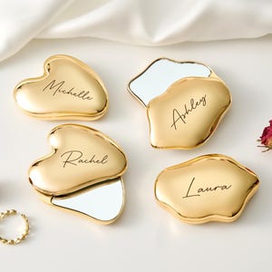 May include: Four gold compact mirrors with personalized names: Michelle, Ashley, Rachel, and Laura. Two are heart-shaped, and two are irregularly shaped. Each mirror has a reflective surface. A gold ring and dried roses are also present.