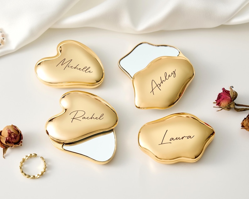 May include: Four gold compact mirrors with personalized names: Michelle, Ashley, Rachel, and Laura. Two are heart-shaped, and two are irregularly shaped. Each mirror has a reflective surface. A gold ring and dried roses are also present.