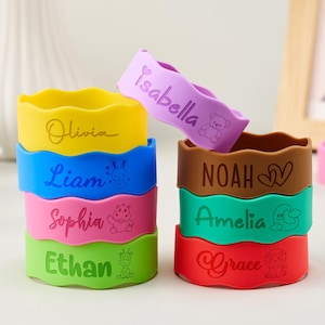 May include: A collection of colorful silicone wristbands with wavy edges, each featuring a unique name and a small animal or heart icon. The bracelets are in yellow, blue, pink, green, purple, brown, teal, and red, displaying names like Olivia, Liam, Sophia, Ethan, Isabella, Noah, Amelia, and Grace.