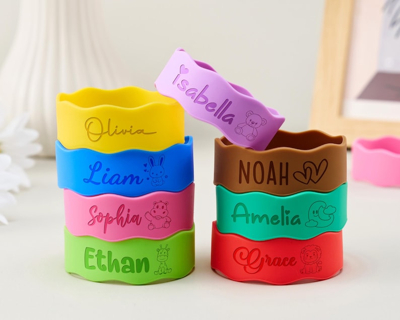 May include: A collection of colorful silicone wristbands with wavy edges, each featuring a unique name and a small animal or heart icon. The bracelets are in yellow, blue, pink, green, purple, brown, teal, and red, displaying names like Olivia, Liam, Sophia, Ethan, Isabella, Noah, Amelia, and Grace.