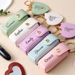 Può includere: Una collezione di astucci per rossetti personalizzati in vari colori pastello, ognuno con un nome stampato sul davanti. Sono visibili anche portachiavi a forma di cuore con disegni floreali. Un rossetto nero e uno specchio a forma di cuore sono anch'essi nell'immagine.
