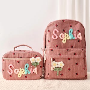 May include: A dusty rose corduroy backpack and matching lunch bag, both featuring a repeating strawberry pattern. Each item is personalized with the name Sophia in multi-colored yarn lettering and decorated with a white crocheted flower applique.