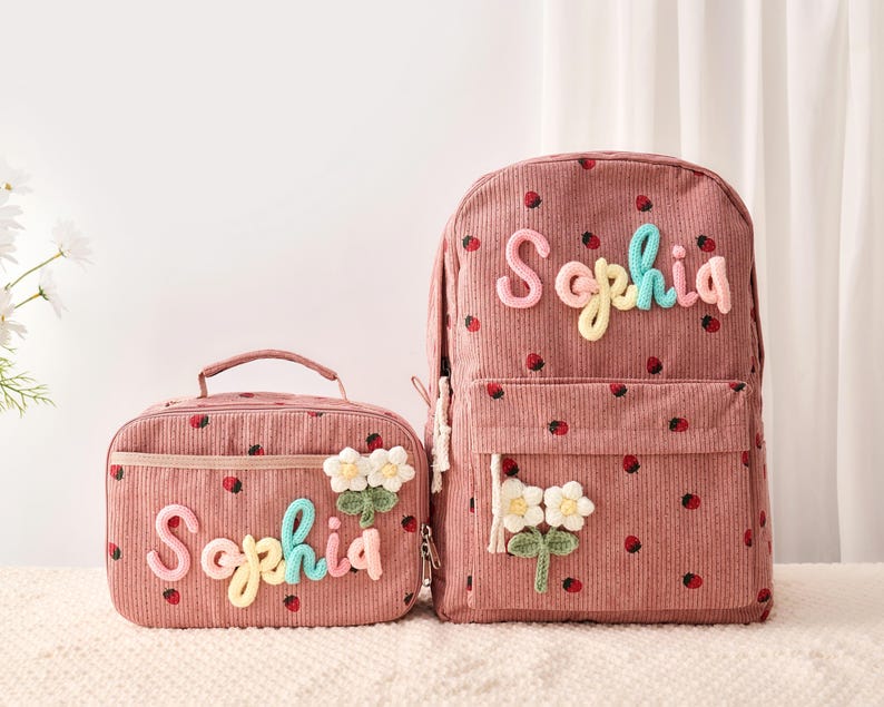 May include: A dusty rose corduroy backpack and matching lunch bag, both featuring a repeating strawberry pattern. Each item is personalized with the name Sophia in multi-colored yarn lettering and decorated with a white crocheted flower applique.