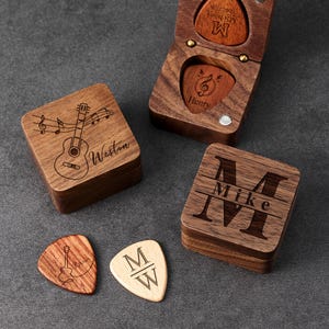 Può includere: Scatole e plettri per chitarra in legno. Una scatola ha una chitarra e note musicali incise sul coperchio, un'altra ha la lettera "M" e il nome "Mike" incisi sul coperchio. Sono visibili due plettri per chitarra.