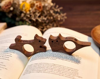 May include: Two wooden cat and dog-shaped book page holders. The cat-shaped holder has the name "Aurora" engraved on it, and the dog-shaped holder has the name "Olivia" engraved on it. Both are resting on an open book.