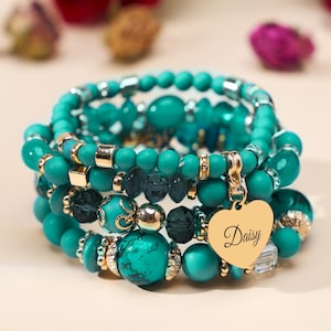 May include: Stack of turquoise beaded bracelets with gold accents and a heart-shaped charm engraved with the name "Daisy". The bracelets feature various bead sizes and textures, creating a layered look.
