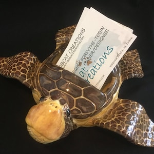 Puede incluir: Un tarjetero de cerámica con forma de tortuga. La tortuga es marrón y beige con un acabado brillante. El tarjetero está lleno de tarjetas de visita blancas. El texto en las tarjetas de visita dice "BOBCAT CREATIONS" y "EGINA FORSYTHE-TOBIN OWNER/DESIGNER".