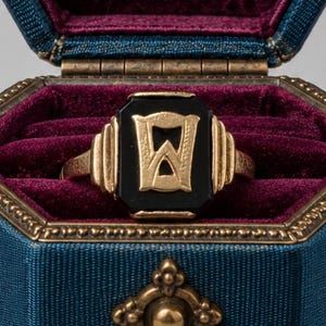 May include: A gold ring featuring a black onyx stone with a gold letter W inlay, presented in a hexagonal blue fabric box with a deep purple velvet interior, ornate brass feet, and a decorative brass latch on the front of the container.