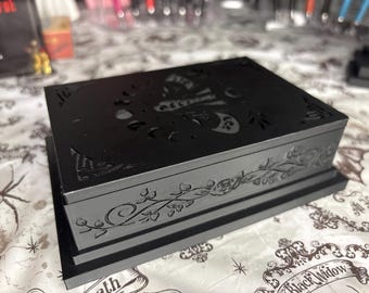 May include: A black rectangular box with a decorative lid featuring a cut-out butterfly and moon design. The sides of the box are adorned with floral and vine patterns. The box sits on a black base.