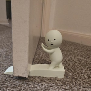May include: A white doorstop shaped like a small, stylized figure with arms outstretched, appearing to hold a door open. The doorstop is positioned to prevent a white door from closing. The figure has a simple face with black dot eyes and a small mouth.