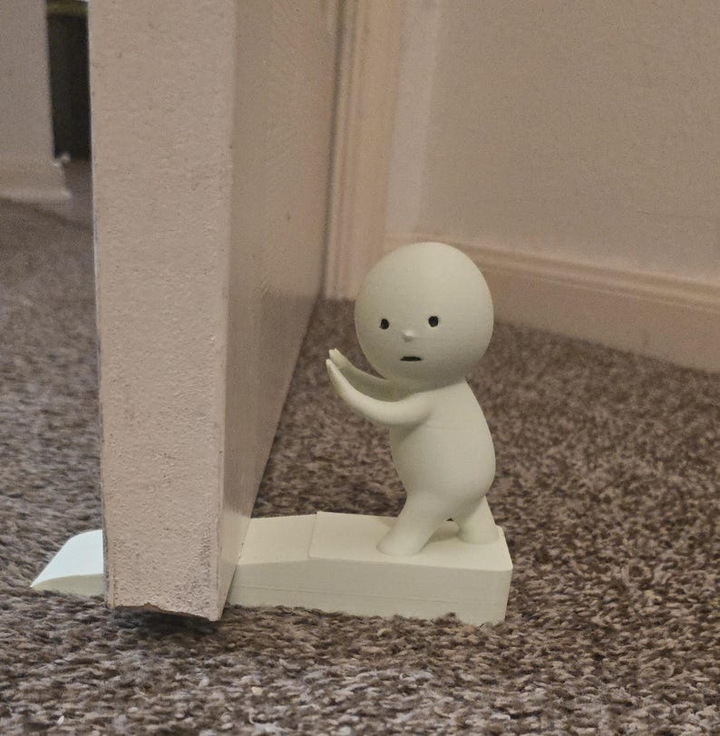 May include: A white doorstop shaped like a small, stylized figure with arms outstretched, appearing to hold a door open. The doorstop is positioned to prevent a white door from closing. The figure has a simple face with black dot eyes and a small mouth.