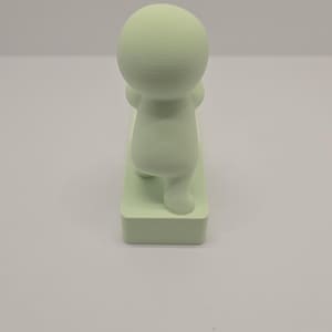 May include: A light green figurine of a person in a yoga pose. The figure has a rounded head and body, with arms raised and legs bent. It is standing on a rectangular base.