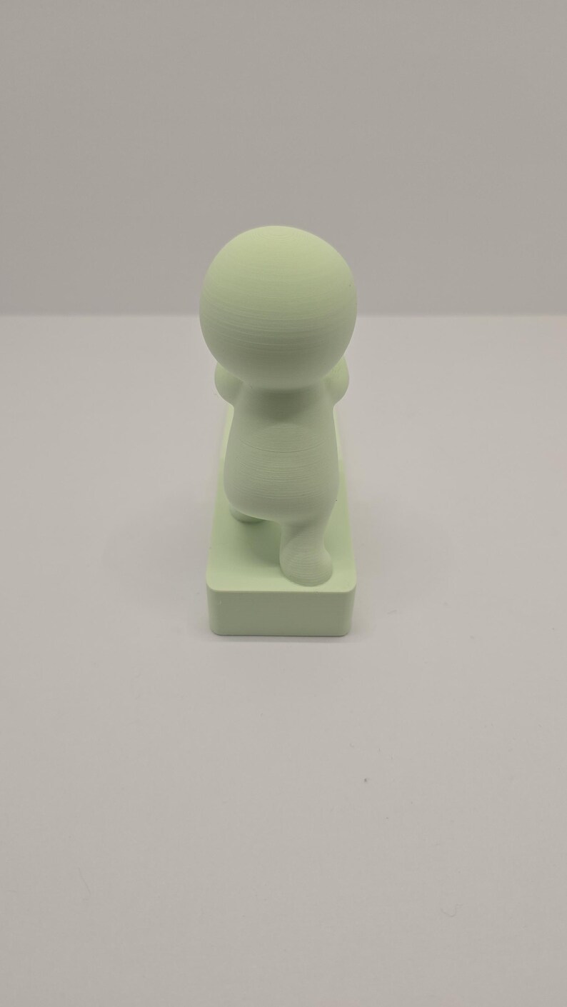 May include: A light green figurine of a person in a yoga pose. The figure has a rounded head and body, with arms raised and legs bent. It is standing on a rectangular base.