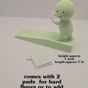 May include: A light green door stop with a small figure on top, approximately 1 inch high and 7 inches long. Includes two pads for hard floors or to add height.