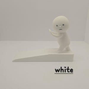 May include: A white figurine of a small person with a round head and outstretched arms, standing on a white platform. The figurine is next to a white doorstop with the word "white" in black text.