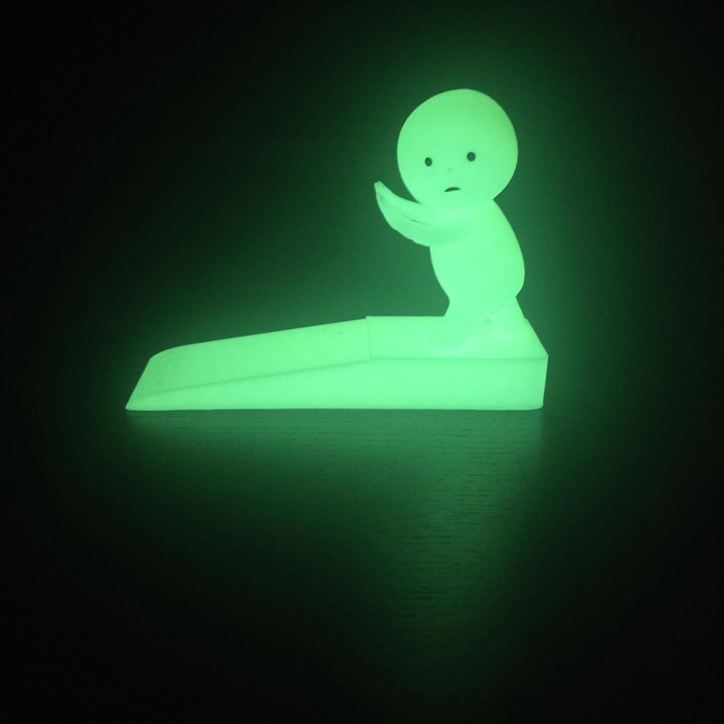 May include: A glowing green doorstop in the shape of a stylized figure. The figure has a rounded head, small facial features, and an outstretched arm. The doorstop is rectangular with a slight incline.
