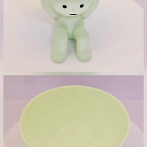 May include: A light green figurine holding a round tray, with jewelry on the tray. The figurine has a minimalist design with black dot eyes and a small mouth. The tray is filled with gold-colored necklaces, rings, and earrings.