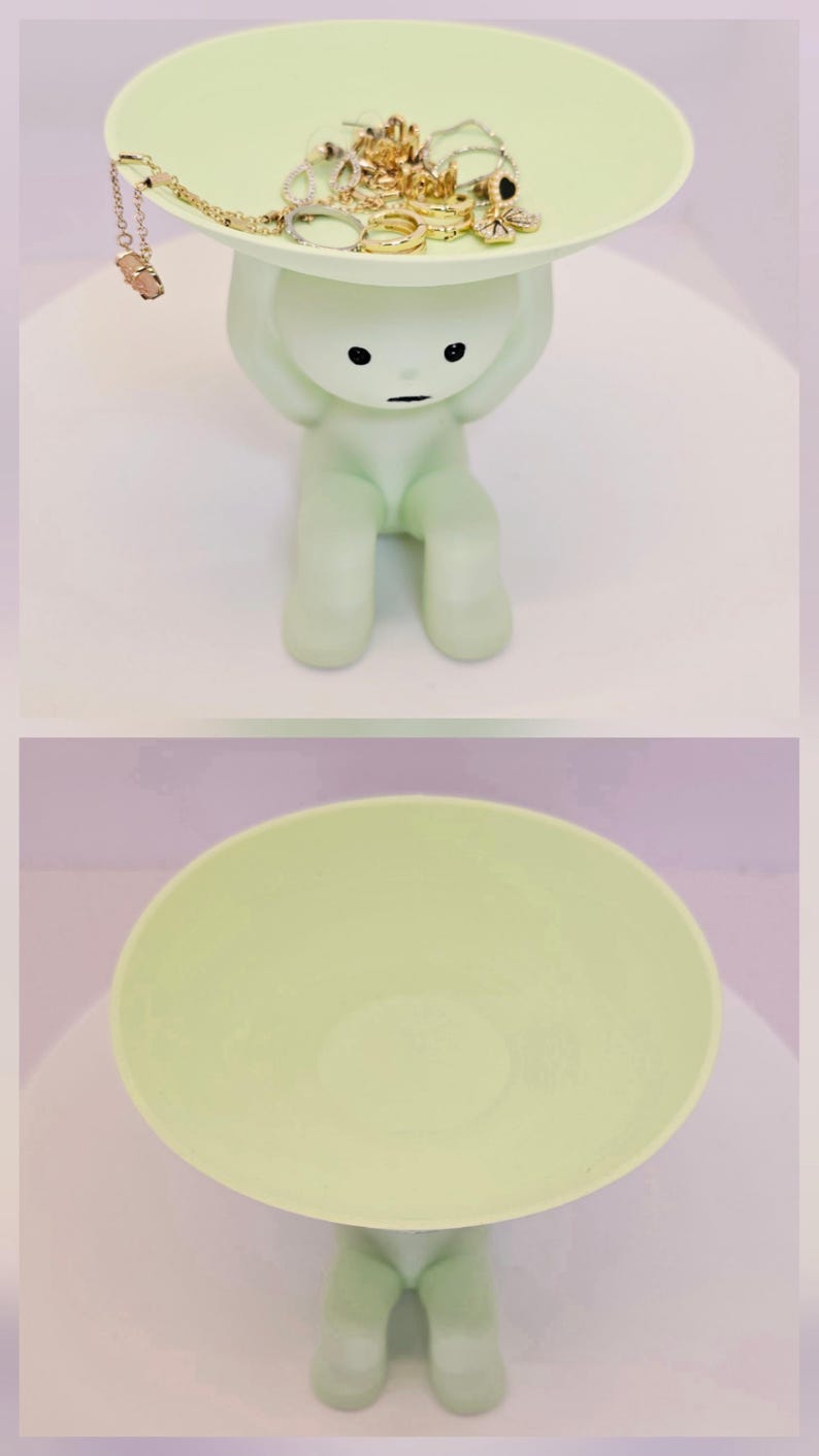 May include: A light green figurine holding a round tray, with jewelry on the tray. The figurine has a minimalist design with black dot eyes and a small mouth. The tray is filled with gold-colored necklaces, rings, and earrings.