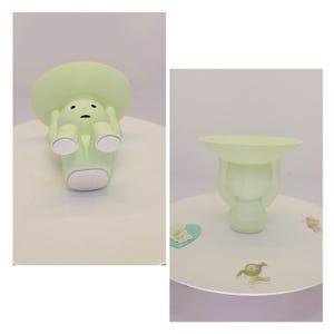 May include: A light green decorative figurine in the shape of a person holding a shallow dish. The figurine has a round head and a sad expression. The dish is held above the head. The figurine is on a white surface.