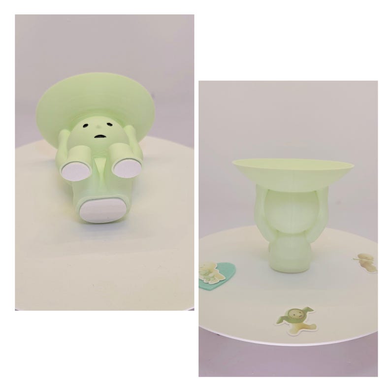 May include: A light green decorative figurine in the shape of a person holding a shallow dish. The figurine has a round head and a sad expression. The dish is held above the head. The figurine is on a white surface.