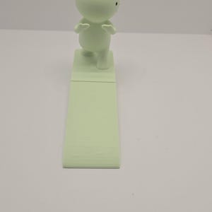 May include: A light green figurine of a small person with arms raised, standing on a rectangular base. The base is connected to a longer, textured, rectangular piece. The figurine has a round head and small black eyes.
