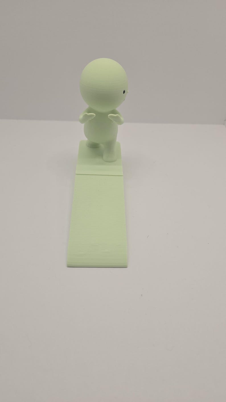May include: A light green figurine of a small person with arms raised, standing on a rectangular base. The base is connected to a longer, textured, rectangular piece. The figurine has a round head and small black eyes.