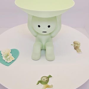 May include: A light green figurine of a person holding a round tray above their head. The figurine has black eyes and a mouth. Small stickers of cartoon figures are scattered around the base.