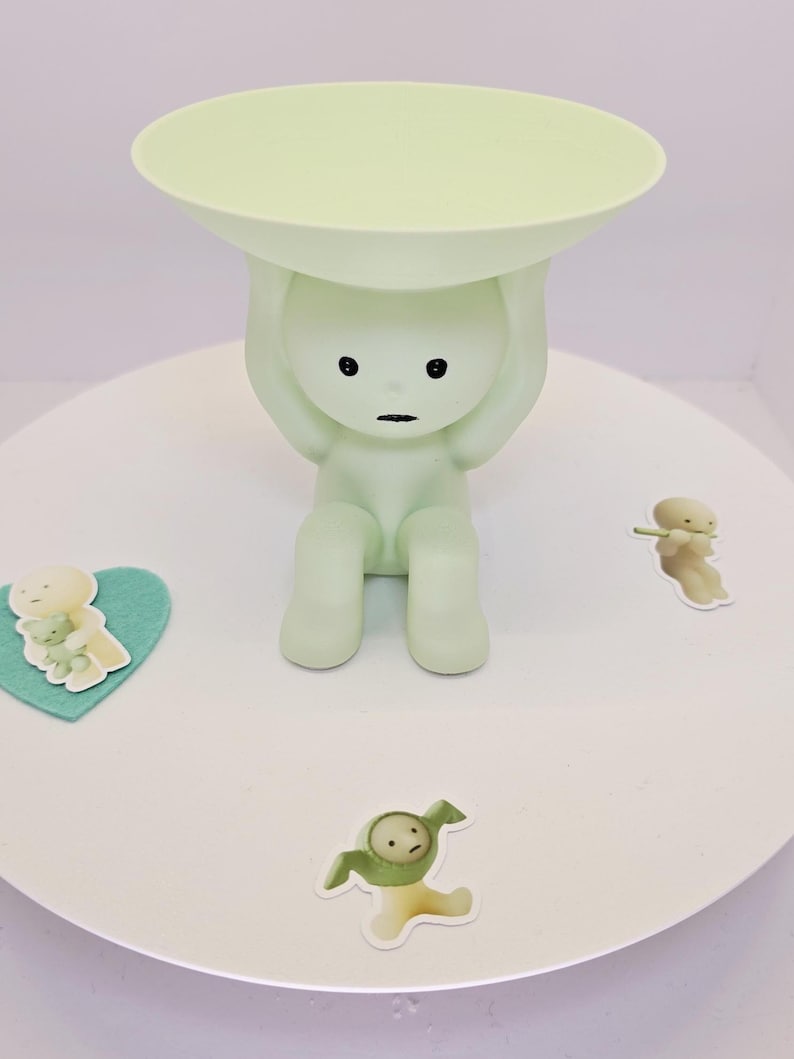 May include: A light green figurine of a person holding a round tray above their head. The figurine has black eyes and a mouth. Small stickers of cartoon figures are scattered around the base.