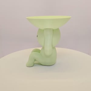 May include: A light green decorative figurine in the shape of a seated figure holding a shallow bowl above its head. The figurine is a solid color and sits on a white surface.