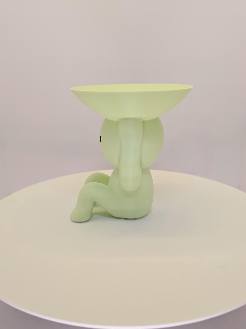 May include: A light green decorative figurine in the shape of a seated figure holding a shallow bowl above its head. The figurine is a solid color and sits on a white surface.
