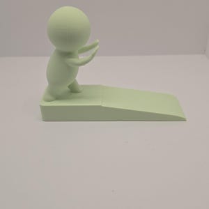 May include: A light green doorstop featuring a stylized figure with arms raised, standing on a rectangular base with a sloped edge. The doorstop is made of a solid, smooth material.