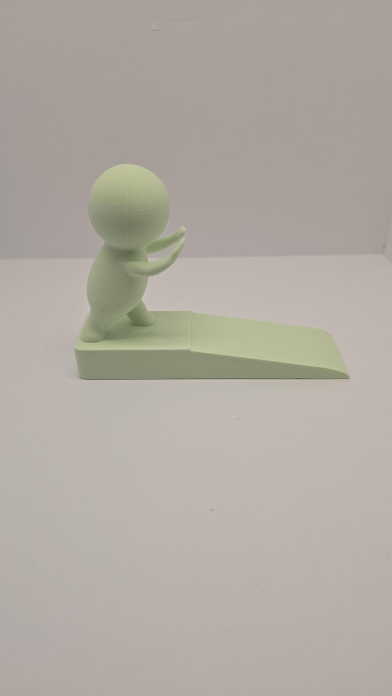 May include: A light green doorstop featuring a stylized figure with arms raised, standing on a rectangular base with a sloped edge. The doorstop is made of a solid, smooth material.