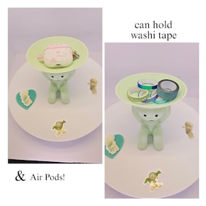 May include: A light green figurine holding a light green plate. The plate holds a pink and white Air Pods case and washi tape. The text says "can hold washi tape" and "& Air Pods!"