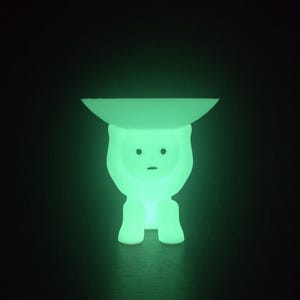 May include: A glowing green decorative lamp in the shape of a stylized figure. The lamp has a flat, circular top and a body with a simple face. The lamp emits a soft, green light against a dark background.