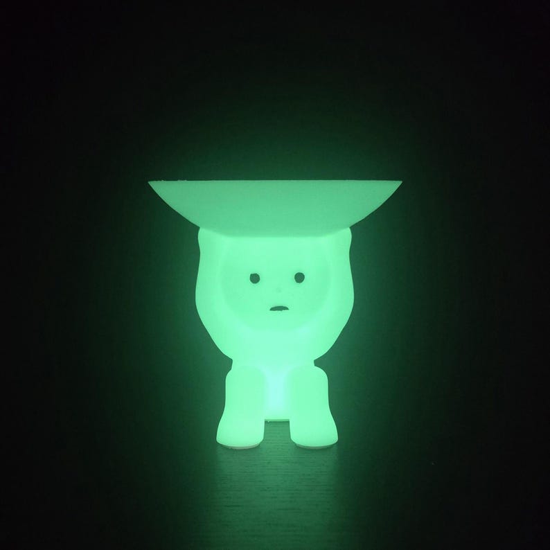 May include: A glowing green decorative lamp in the shape of a stylized figure. The lamp has a flat, circular top and a body with a simple face. The lamp emits a soft, green light against a dark background.