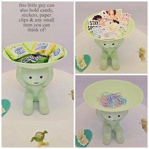 May include: A light green figurine holding a round tray. The tray is filled with candy, stickers, and paper clips. The figurine has a minimalist design and is sitting down. Text on the image says, "this little guy can also hold candy, stickers, paper clips & any small item you can think of!"